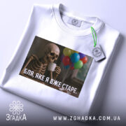 Футболка я вже старе. Біла футболка, складена на столі, з принтом скелета, що тримає чашку, на фоні кольорових повітряних куль.