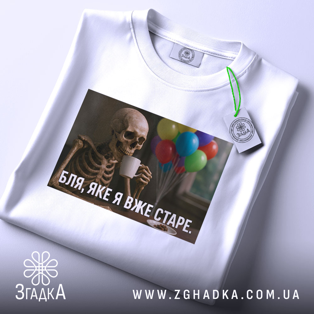 Футболка я вже старе. Біла футболка, складена на столі, з принтом скелета, що тримає чашку, на фоні кольорових повітряних куль.