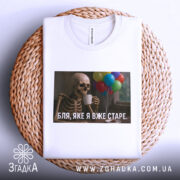 Футболка я вже старе. Біла футболка, складена на плетеному кошику, з принтом скелета, що тримає чашку, на фоні кольорових повітряних куль.