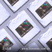 Футболка я вже старе. Біла футболка, розкладена на поверхні, з принтом скелета, що тримає чашку, на фоні кольорових повітряних куль.