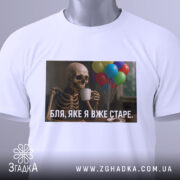 Футболка я вже старе. Біла футболка на манекені, з принтом скелета, що тримає чашку, на фоні кольорових повітряних куль.