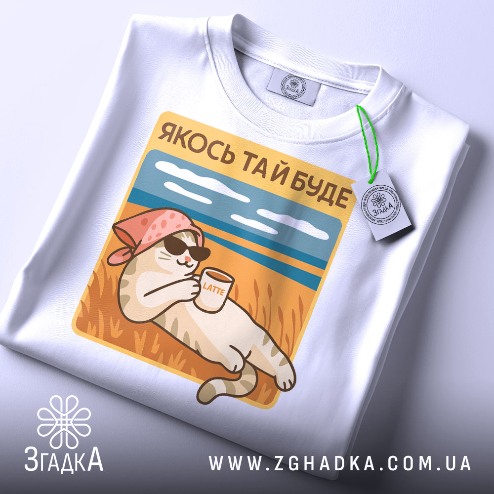 Футболка кіт якось буде. Складена біла футболка на світлому фоні, ракурс зверху. Принт з котом у рожевій бандані, що тримає чашку з написом 'LATTE', на фоні пляжу з блакитним небом.