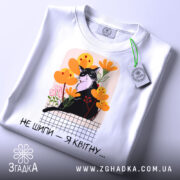 Футболка кіт не шипи, білого кольору, складена на поверхні, фон світло-сірий, принт з чорним котом серед жовтих і рожевих квітів, текст під принтом чорний, на футболці є етикетка з зеленим шнурком.