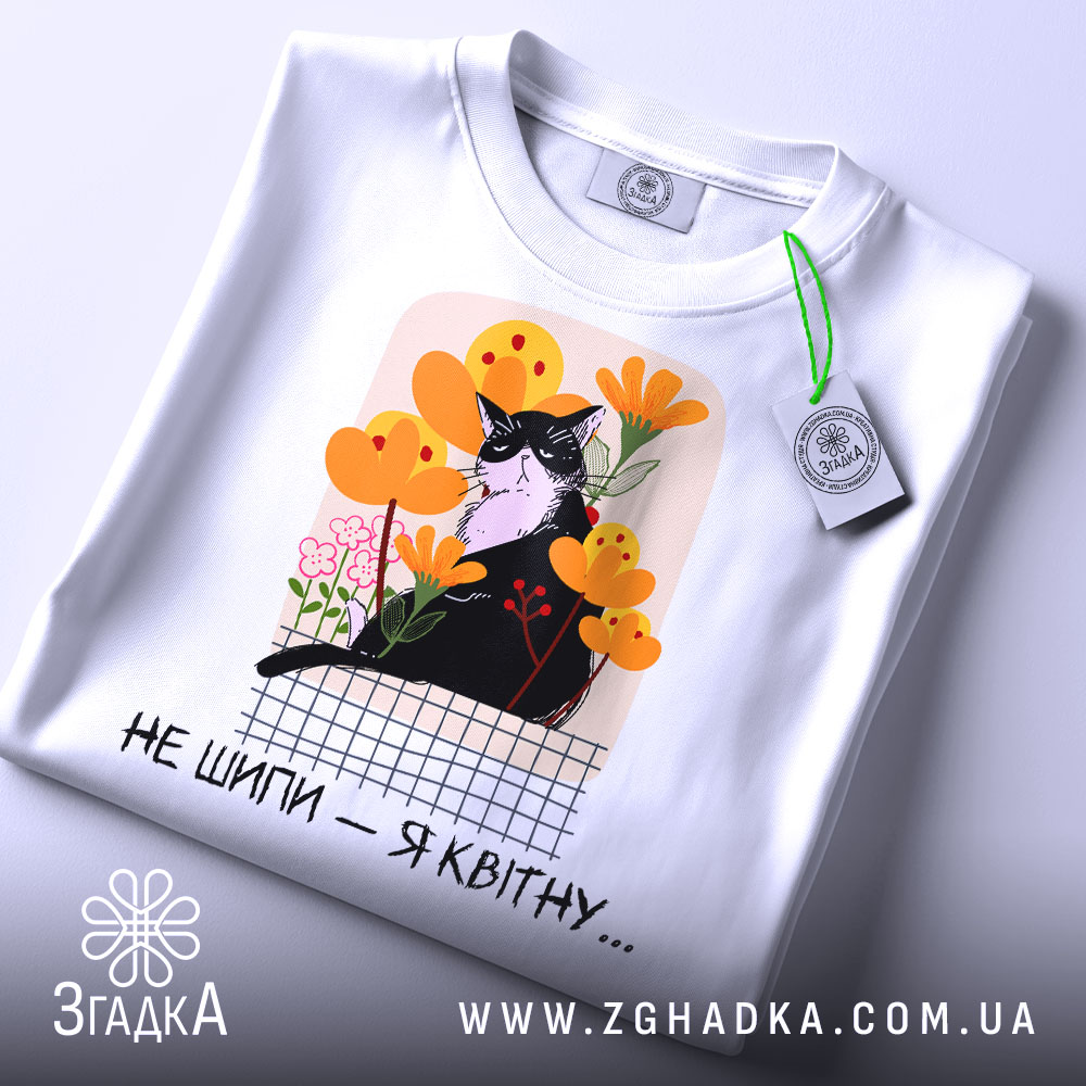 Футболка кіт не шипи, білого кольору, складена на поверхні, фон світло-сірий, принт з чорним котом серед жовтих і рожевих квітів, текст під принтом чорний, на футболці є етикетка з зеленим шнурком.