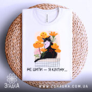 Футболка кіт не шипи, білого кольору, складена на плетеній кошику, фон білий, принт з чорним котом серед жовтих і рожевих квітів, текст під принтом чорний.