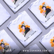 Футболка кіт не шипи, білого кольору, розкладена на поверхні, фон світло-сірий, кілька футболок з однаковим принтом, котом на фоні жовтих і рожевих квітів, текст під принтом чорний.