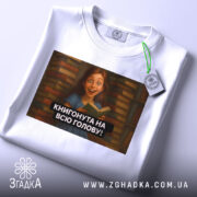 Футболка книгонута на всю голову, білого кольору, з ракурсу зверху, на фоні світлого кольору, з принтом, що зображує дівчину з книгою на фоні книжкових полиць, з етикеткою зеленого кольору.