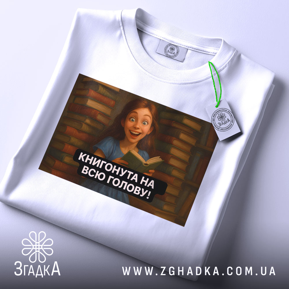 Футболка книгонута на всю голову, білого кольору, з ракурсу зверху, на фоні світлого кольору, з принтом, що зображує дівчину з книгою на фоні книжкових полиць, з етикеткою зеленого кольору.