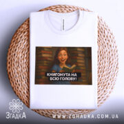 Футболка книгонута на всю голову, білого кольору, з ракурсу зверху, на фоні плетеного кошика, з принтом, що зображує дівчину з книгою на фоні книжкових полиць.