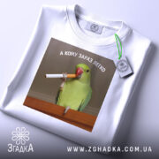 Футболка кому зараз легко, біла, зображена зверху, фон світлий, принт з папугою, що тримає сигарету, текст зверху: 'А КОМУ ЗАРАЗ ЛЕГКО', з етикеткою на рукаві.