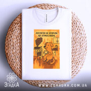 Футболка котлети не згоріли. Біла футболка з принтом, на якому зображений жовтий собака, що сидить на стільці з чашкою в руках. На фоні видно кухонну плиту з димом та вогнем. Футболка зображена на плетеній кошику.