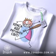 Футболка краса рятує світ. Біла футболка, складена на столі, з принтом, що зображає персонажа з короною і битою. На футболці є етикетка з зеленим шнурком, фон світлий.