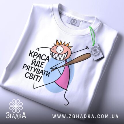 Футболка краса рятує світ. Біла футболка, складена на столі, з принтом, що зображає персонажа з короною і битою. На футболці є етикетка з зеленим шнурком, фон світлий.