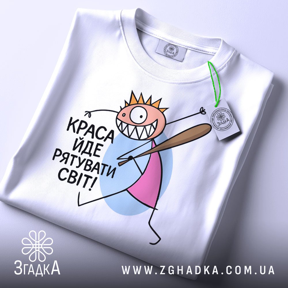 Футболка краса рятує світ. Біла футболка, складена на столі, з принтом, що зображає персонажа з короною і битою. На футболці є етикетка з зеленим шнурком, фон світлий.