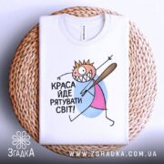 Футболка краса рятує світ. Біла футболка, складена на плетеній кошику, з принтом, що зображає персонажа з короною і битою. Фон світлий, текстура кошика помітна.