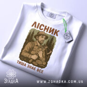 Футболка лісник тиша знає, біла футболка складена, ракурс зверху, фон світлий, принт з лісником та текстом, тег на комірці.