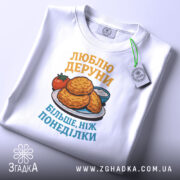 Футболка люблю деруни подарунок, біла, складена на столі, фон світло-сірий, принт з картинкою дерунів на тарілці з помідором і соусом, текст жовтим і блакитним кольором, з етикеткою.