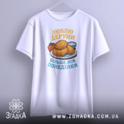 Футболка люблю деруни подарунок, біла, зображена з фронтального ракурсу, фон світло-сірий, принт з картинкою дерунів на тарілці з помідором і соусом, текст жовтим і блакитним кольором.