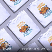 Футболка люблю деруни подарунок, біла, складена в хаотичному порядку, фон світло-сірий, принт з картинкою дерунів на тарілці з помідором і соусом, текст жовтим і блакитним кольором.