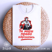 Футболка маєш право відрізнятись. Білий колір, зображена зверху, фон світлий, принт з червоним текстом і чорною ілюстрацією жінки, розташована на плетеній основі.