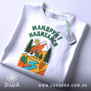 Футболка мандруй і надихайся, біла, з верхнього ракурсу, на столі, фон світло-сірий, принт з гірським пейзажем, річкою та деревами, кольори яскраві: червоний, зелений, жовтий, блакитний, з етикеткою.