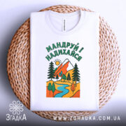 Футболка мандруй і надихайся, біла, з верхнього ракурсу, на плетеному кошику, фон білий, принт з гірським пейзажем, річкою та деревами, кольори яскраві: червоний, зелений, жовтий, блакитний.