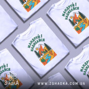 Футболка мандруй і надихайся, біла, з верхнього ракурсу, кілька футболок на столі, фон світло-сірий, принт з гірським пейзажем, річкою та деревами, кольори яскраві: червоний, зелений, жовтий, блакитний.