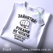 Футболка моя вас зафігачить. Біла футболка, складена на поверхні, з видимим круглим вирізом. На футболці великий принт з кулаком і текстом, з зеленим ярликом, що звисає з коміра.