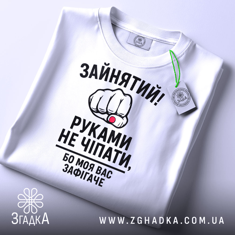 Футболка моя вас зафігачить. Біла футболка, складена на поверхні, з видимим круглим вирізом. На футболці великий принт з кулаком і текстом, з зеленим ярликом, що звисає з коміра.