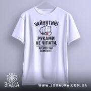 Футболка моя вас зафігачить. Біла футболка з коротким рукавом, підвішена на стіні, з фронтального ракурсу. Принт з кулаком і текстом розташований по центру.