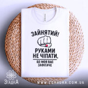 Футболка моя вас зафігачить. Біла футболка, складена на плетеній кошику, з видимим круглим вирізом. Принт з кулаком і текстом розташований по центру.