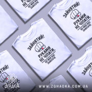 Футболка моя вас зафігачить. Білі футболки, складені в хаотичному порядку на фоні світло-сірого кольору. Кожна футболка має принт з кулаком і текстом, що повторюється.