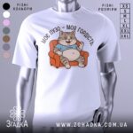 Футболка моє пузо гордість. Біла футболка з принтом кота, що сидить на дивані, з ракурсу спереду. Фон світло-сірий, принт кольоровий, котик у блакитній футболці з усмішкою і піднятою рукою.