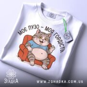 Футболка моє пузо гордість. Біла футболка, складена на поверхні, з принтом кота на дивані. Фон світло-сірий, на футболці видно етикетку з зеленим шнурком.