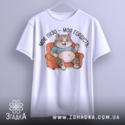 Футболка моє пузо гордість. Біла футболка з принтом кота, що сидить на дивані, з ракурсу спереду. Фон світло-сірий, принт кольоровий, котик у блакитній футболці з усмішкою.