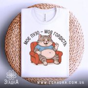 Футболка моє пузо гордість. Біла футболка, складена на плетеній основі, з принтом кота на дивані. Фон світло-сірий, принт кольоровий, котик усміхається і підняв руку.