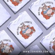 Футболка моє пузо гордість. Біла футболка з принтом кота, що сидить на дивані, з ракурсу зверху. Фон світло-сірий, принт кольоровий, котик у блакитній футболці з усмішкою.