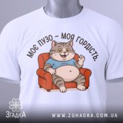 Футболка моє пузо гордість. Біла футболка з принтом кота, що сидить на дивані, з ракурсу спереду. Фон світло-сірий, принт кольоровий, котик у блакитній футболці з усмішкою і піднятою рукою.
