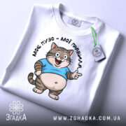 Футболка моє пузо мої правила. Біла футболка, складена на столі, з принтом кота в блакитній футболці, що усміхається. На футболці є етикетка з зеленим шнурком. Фон світло-сірий, деталі принта чітко видимі.