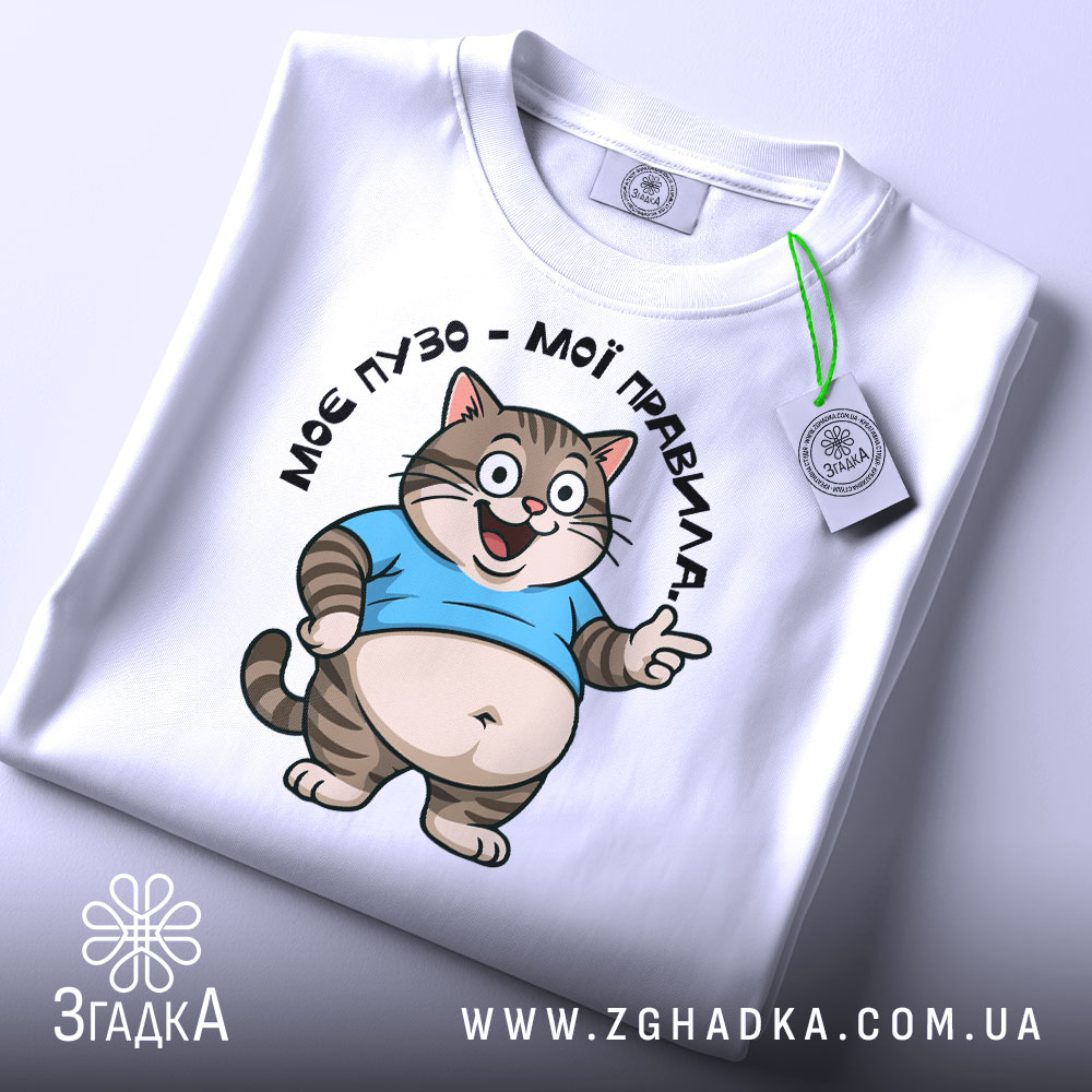 Футболка моє пузо мої правила. Біла футболка, складена на столі, з принтом кота в блакитній футболці, що усміхається. На футболці є етикетка з зеленим шнурком. Фон світло-сірий, деталі принта чітко видимі.