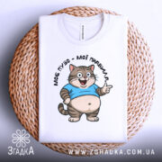 Футболка моє пузо мої правила. Біла футболка, складена на кошику з плетеного матеріалу, з принтом кота в блакитній футболці, що усміхається. Фон світло-сірий, деталі принта чітко видимі.