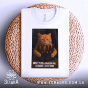 Футболка моє тіло машина. Біла футболка, складена на кошику з плетеного матеріалу, з принтом кота, що тримає пляшки пива. Фон світлий, що підкреслює текст на футболці.