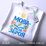 Футболка Мова моя зброя, білого кольору, зображена зверху на плоскій поверхні, фон світло-сірий, принт з синім текстом "МОВА-МОЯ ЗБРОЯ" та елементами книжки, пера і рослин, з етикеткою на комірці.