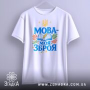 Футболка Мова моя зброя, білого кольору, зображена з близької відстані на вішалці, фон світло-сірий, принт містить синій текст "МОВА-МОЯ ЗБРОЯ" з елементами книжки, пера та рослин.