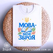 Футболка Мова моя зброя, білого кольору, зображена зверху на плоскій поверхні, фон світло-сірий, принт з синім текстом "МОВА-МОЯ ЗБРОЯ" та елементами книжки, пера і рослин, на плетеній основі.