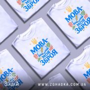Футболка Мова моя зброя, білого кольору, зображена зверху на плоскій поверхні, фон світло-сірий, кілька футболок з принтом "МОВА-МОЯ ЗБРОЯ" розкладені разом, з елементами книжки, пера та рослин.