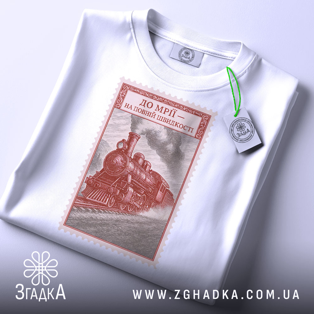 Футболка на повній швидкості, біла, з ракурсу зверху, на фоні світло-сірого, принт з паровозом в червоному кольорі, з етикеткою зеленого кольору.