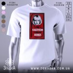 Футболка напис під каблуком, біла, зображена на манекені, фон світло-сірий, принт червоний з чорною обводкою, на якому зображена жінка з коротким волоссям та написом 'ПІД КАБЛУКОМ У СИЛЬНОЇ ЖІНКИ'.