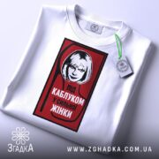 Футболка напис під каблуком, біла, складена на столі, фон світло-сірий, принт червоний з чорною обводкою, на якому зображена жінка з коротким волоссям та написом 'ПІД КАБЛУКОМ У СИЛЬНОЇ ЖІНКИ', з біркою на рукаві.