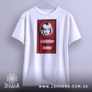 Футболка напис під каблуком, біла, підвішена на вішалці, фон світло-сірий, принт червоний з чорною обводкою, на якому зображена жінка з коротким волоссям та написом 'ПІД КАБЛУКОМ У СИЛЬНОЇ ЖІНКИ'.